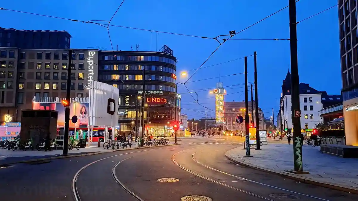 A city street at dusk with tram tracks, illuminated buildings including 'Byporten' and 'Lindex', and traffic lights. The…