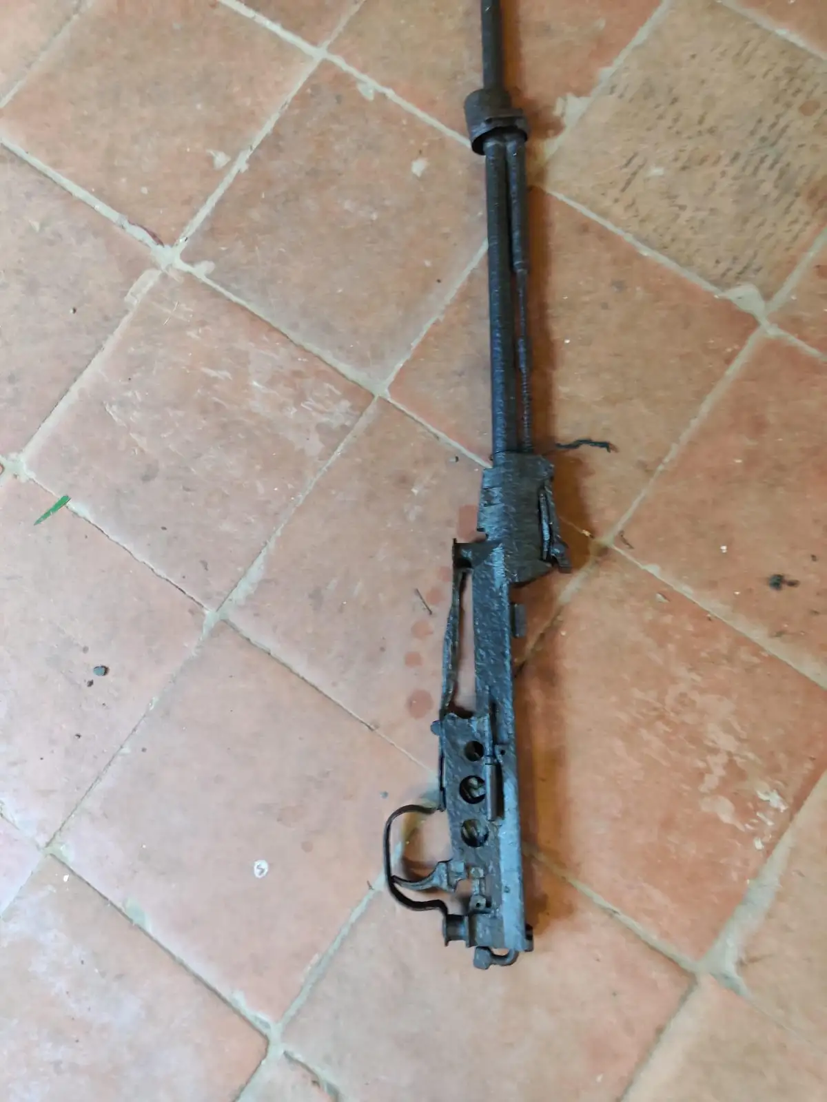 A rusted, stripped-down firearm receiver and barrel assembly lies on a terracotta tiled floor. The overlaid text reads 'i…