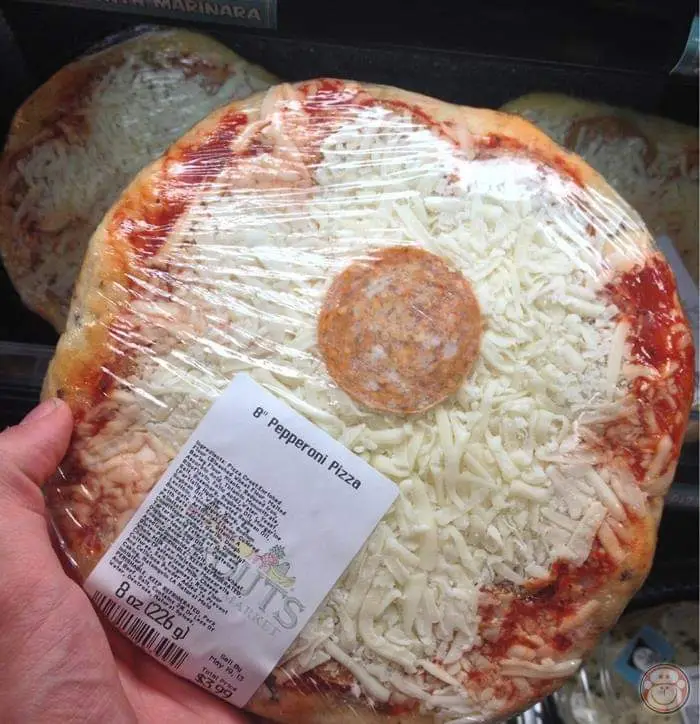 A hand holds an 8-inch pepperoni pizza wrapped in clear plastic wrap, showing a single pepperoni slice in the center and…