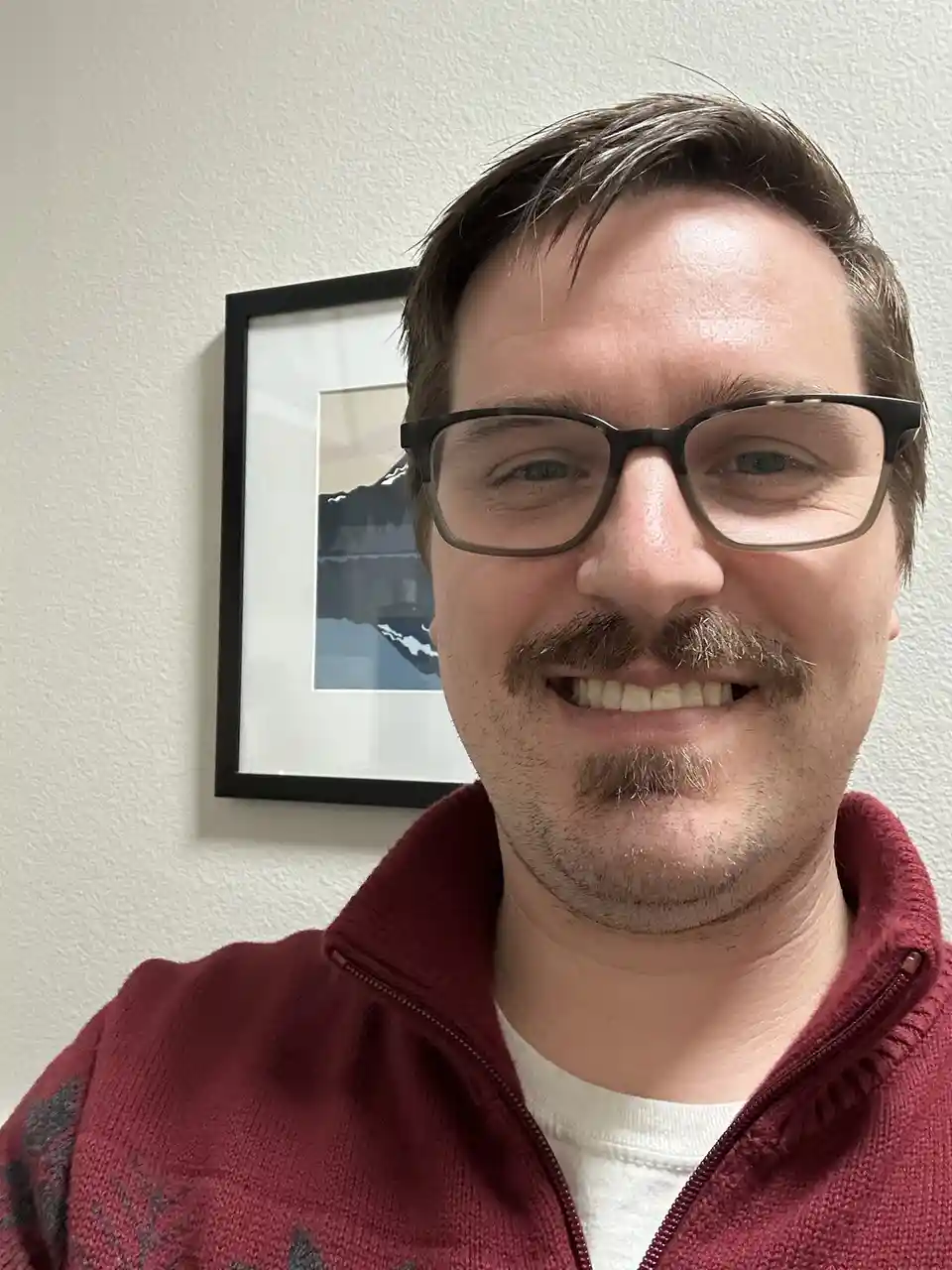 A man with glasses and a prominent mustache smiles at the camera while wearing a red zip-up sweater over a white shirt…