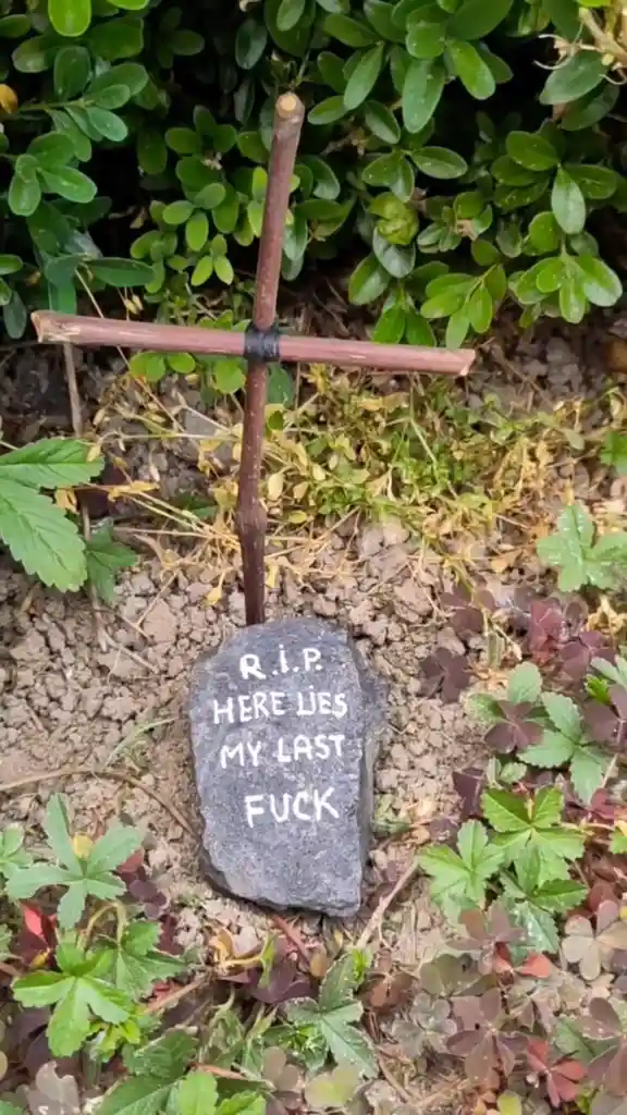 A small, handmade grave marker made of a rock and a twig cross is planted in dirt surrounded by green and reddish clover…