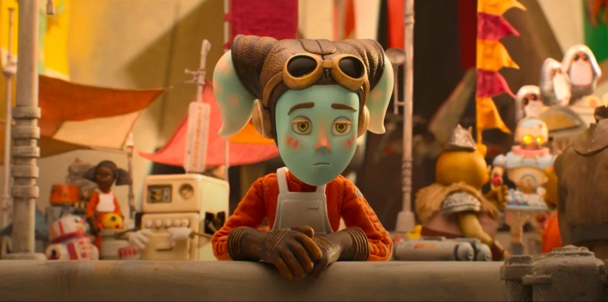 A blue-skinned animated character with large ears, goggles on their head, and a worried expression leans on a counter in a…
