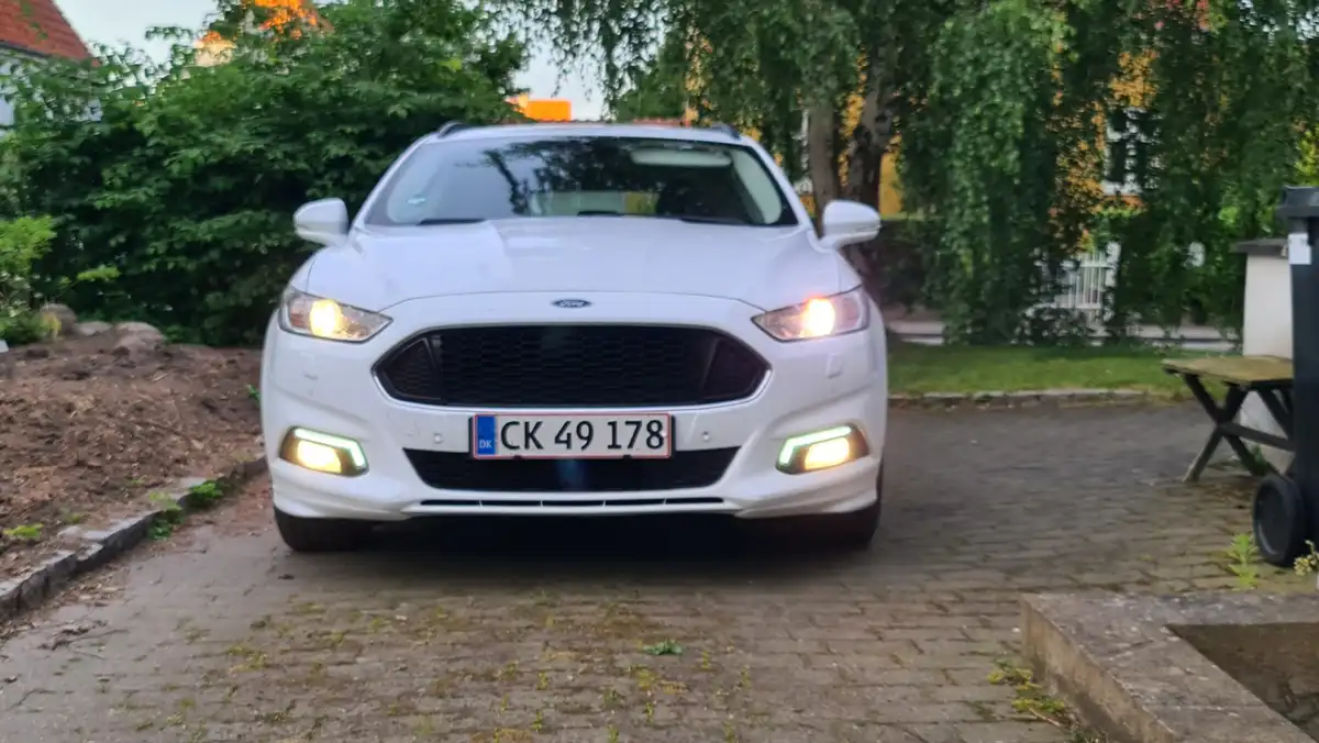 A white Ford car is parked on a paved driveway with its headlights and daytime running lights on. The license plate reads…