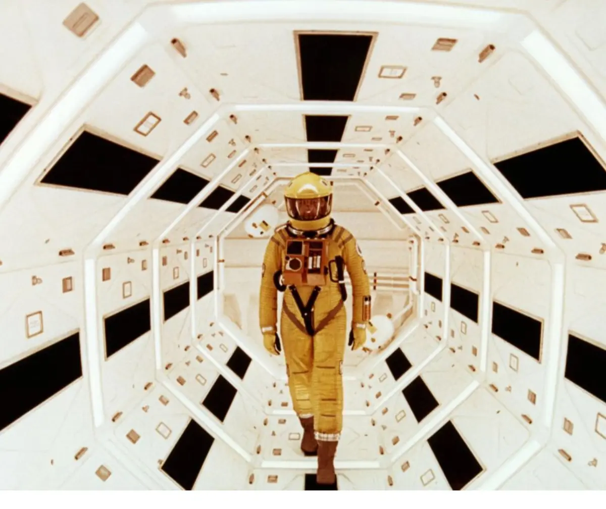 A person in a yellow spacesuit walks down a long, white, hexagonal spaceship corridor with black rectangular panels on the…