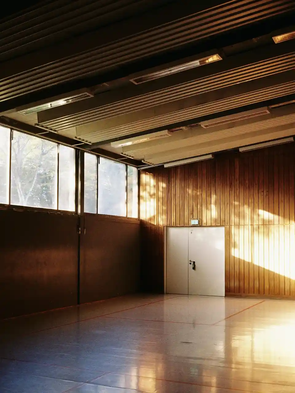 An empty gymnasium with wooden walls, a white door, and large windows letting in sunlight that casts long shadows. The floor…
