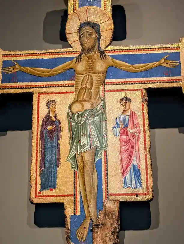 A close-up of a medieval mosaic crucifix depicting Jesus Christ on the cross with two figures below, one in blue and one in…