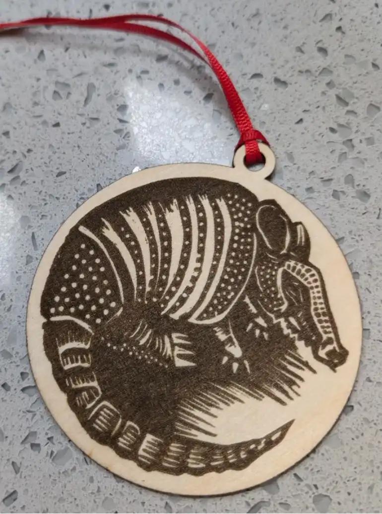 A circular wooden ornament with a laser-etched image of an armadillo, featuring detailed patterns on its shell and tail…