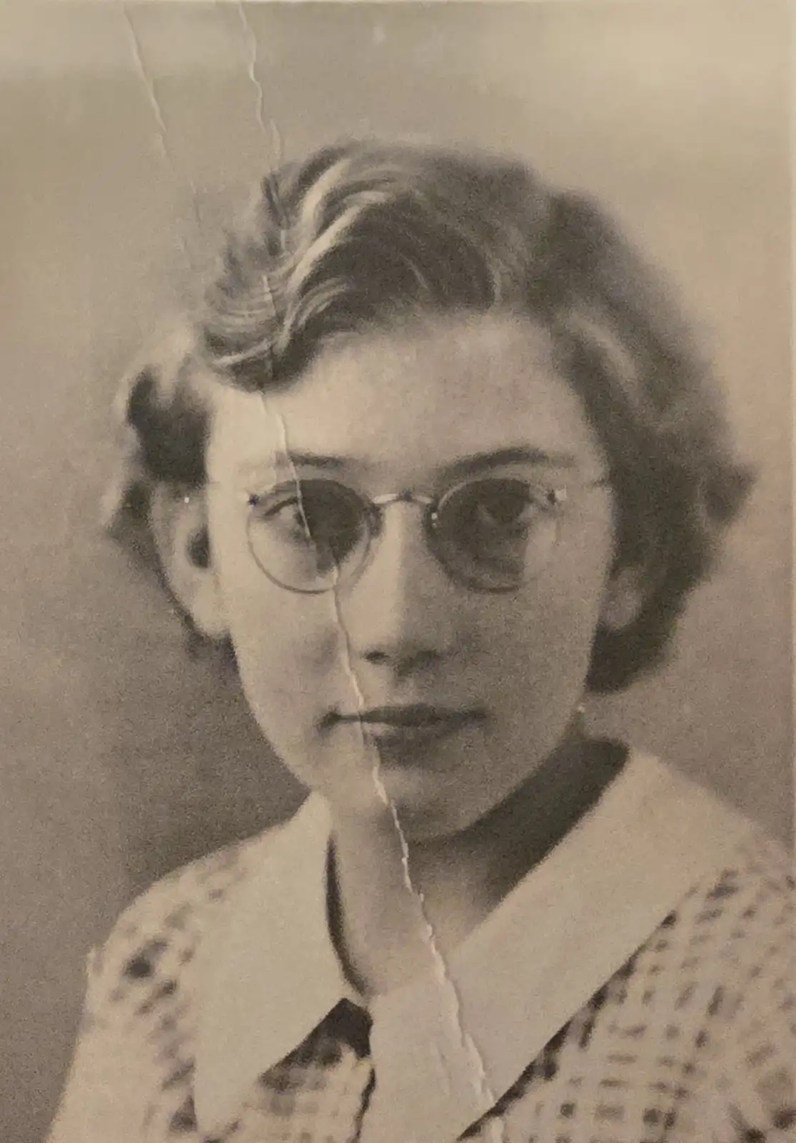 A vintage black-and-white portrait of a young woman with short, wavy hair and round glasses, wearing a collared shirt with a…