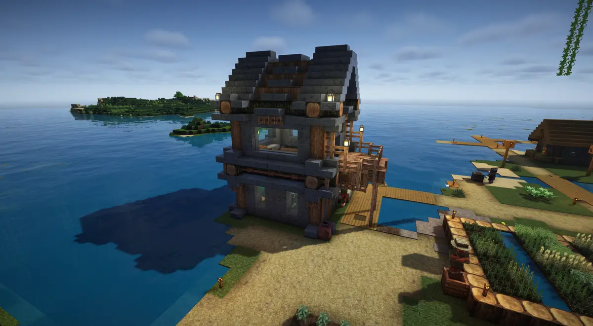 A screenshot from the game Minecraft shows a large, multi-level stone and wood house built on a sandy shore next to a vast…