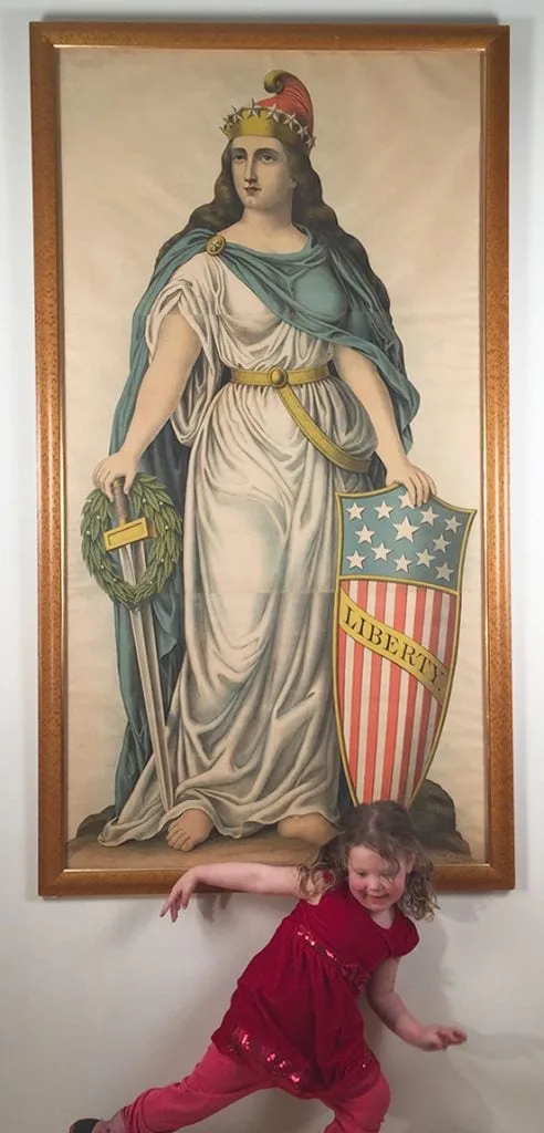 A young girl in a red outfit is playfully posing in front of a large framed painting of Lady Liberty, who holds a shield…