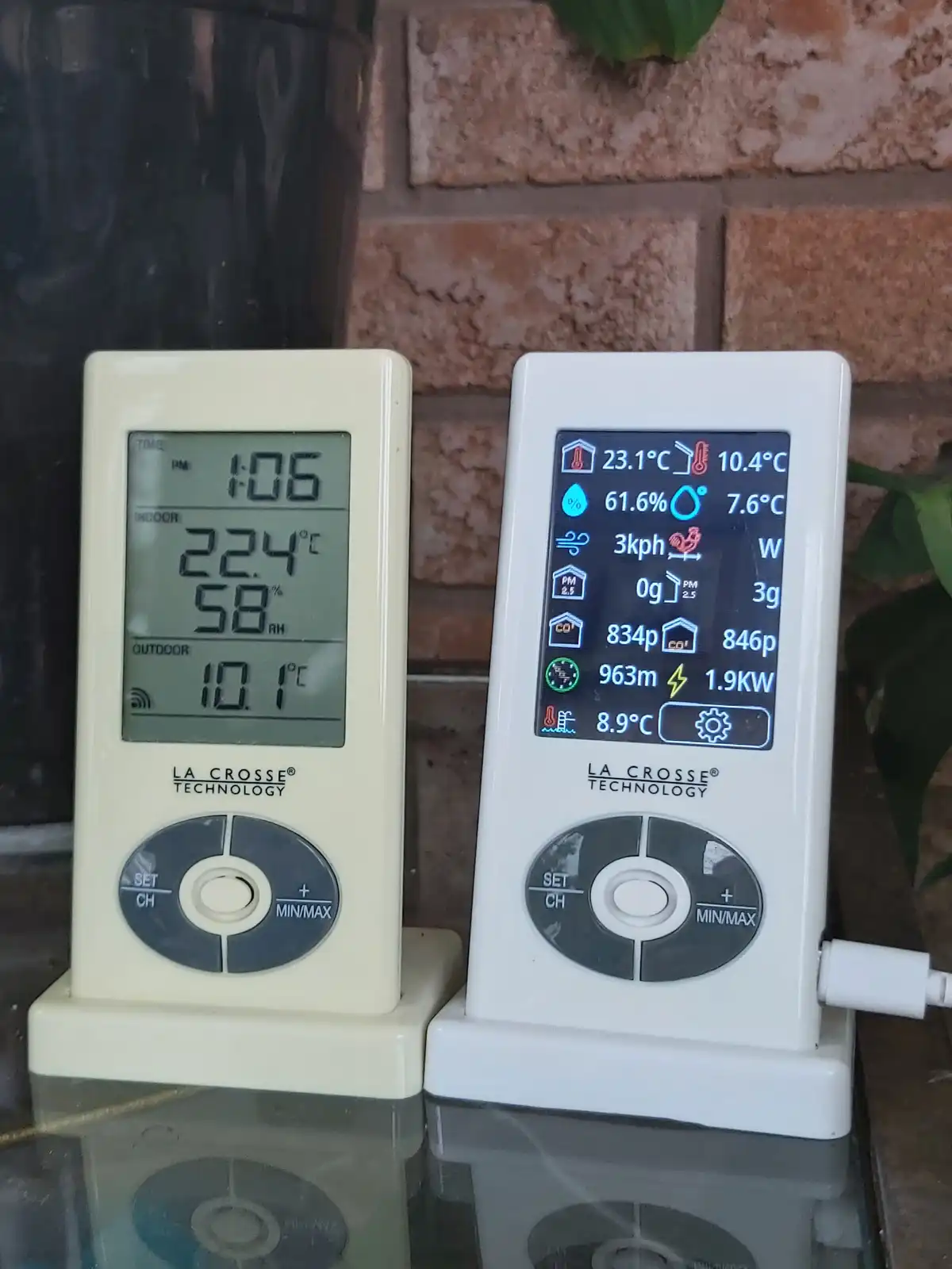 Two La Crosse Technology weather stations sit side by side on a glass surface against a brick wall; the left unit shows…