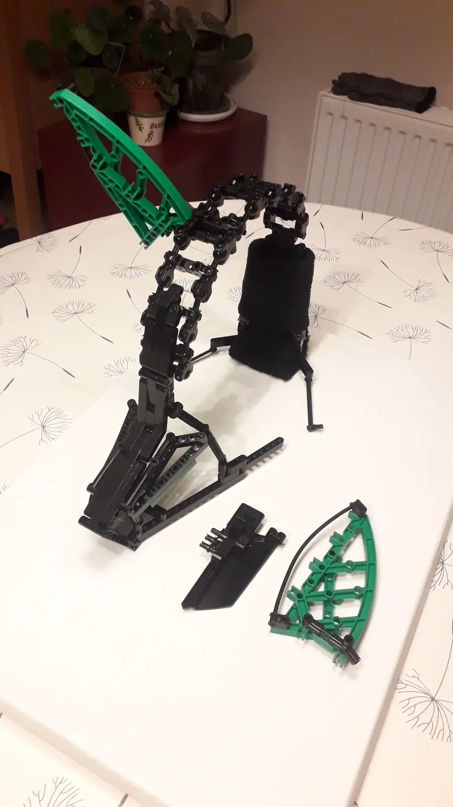 A partially assembled black and green Lego dragon model sits on a white table with dandelion-patterned cloth, its left wing…