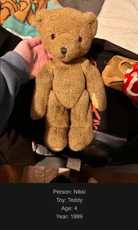 A person's hand holds a worn, brown teddy bear with simple black button eyes and nose, sitting upright on a black bag. The…