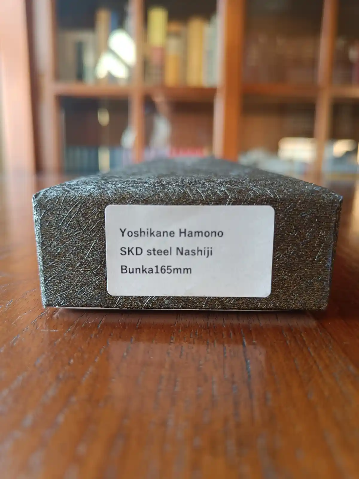 A close-up photo shows a rectangular, textured metal block resting on a wooden surface with a blurred bookshelf in the…