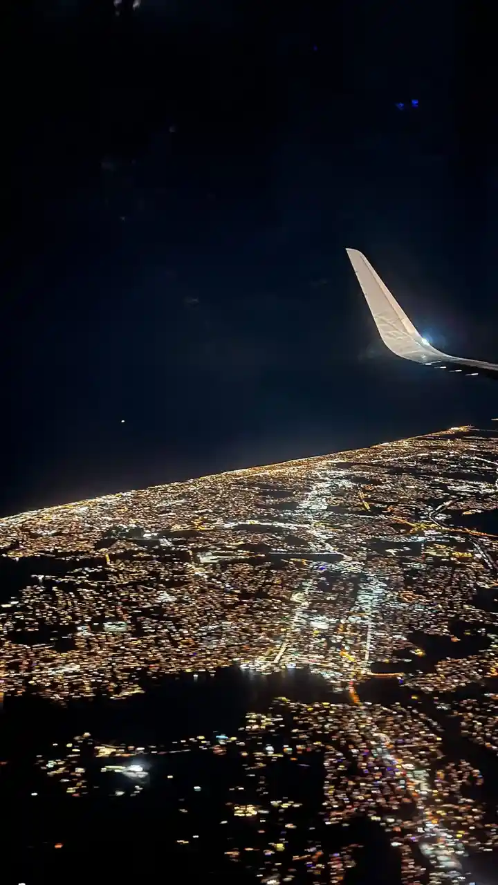 A nighttime aerial view from an airplane window showing the wing and a sprawling cityscape illuminated by countless lights…