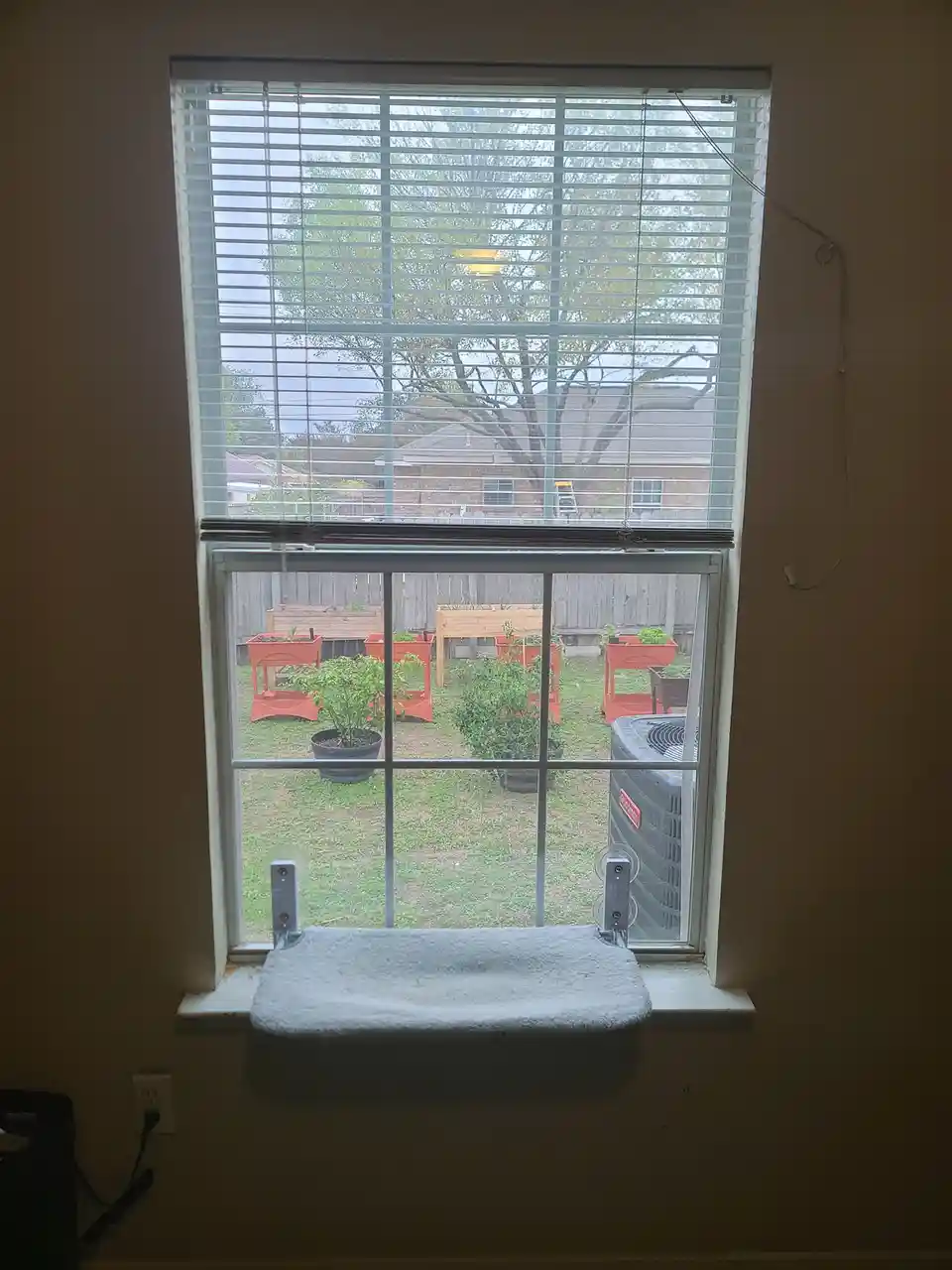 A window with blinds partially open looks out onto a backyard with several orange planters and potted plants. A gray towel…