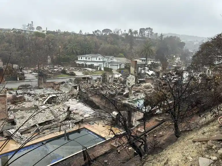 A wide-angle photo shows a neighborhood devastated by fire, with charred trees, rubble, and destroyed homes, including a…