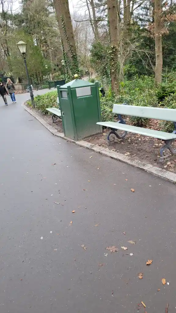 A photo of a park scene with a green trash can and benches next to a paved path, with people walking in the background. The…
