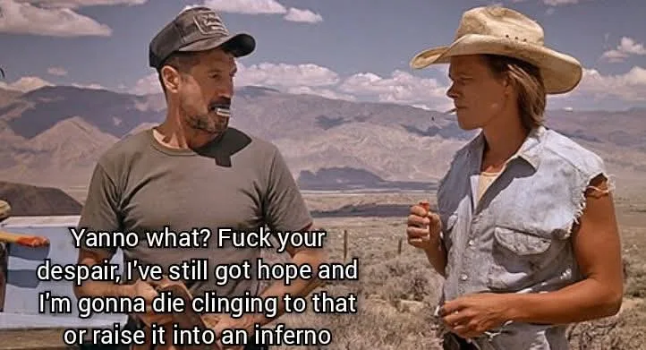 Two characters from a Western film stand in a desert landscape with mountains in the background. The man on the left…