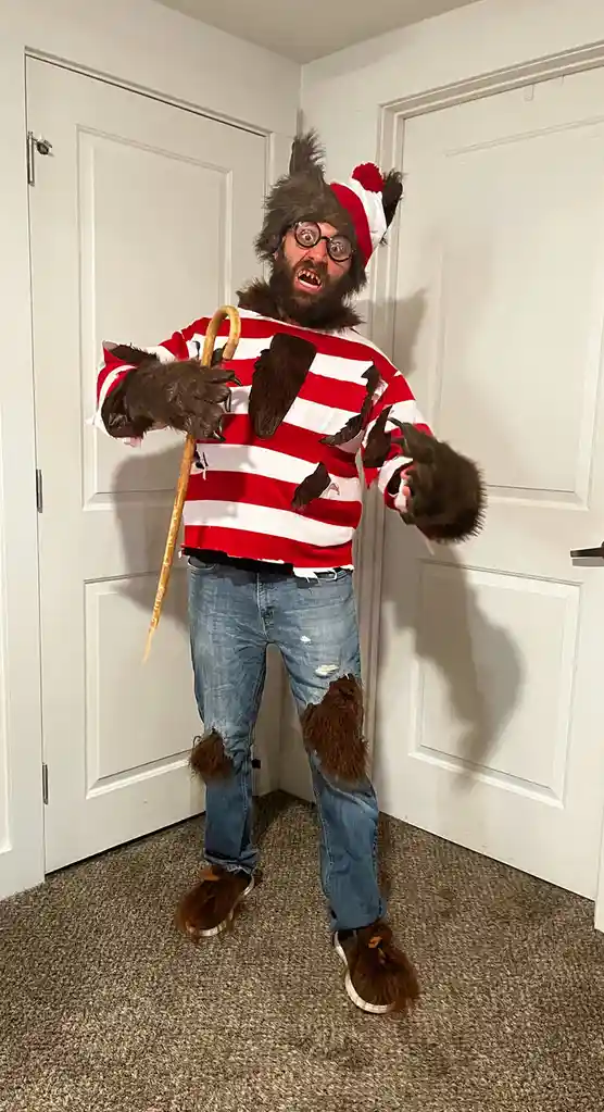 A man in a full-body wolf costume, complete with furry ears, claws, and a red-and-white striped shirt with torn holes…