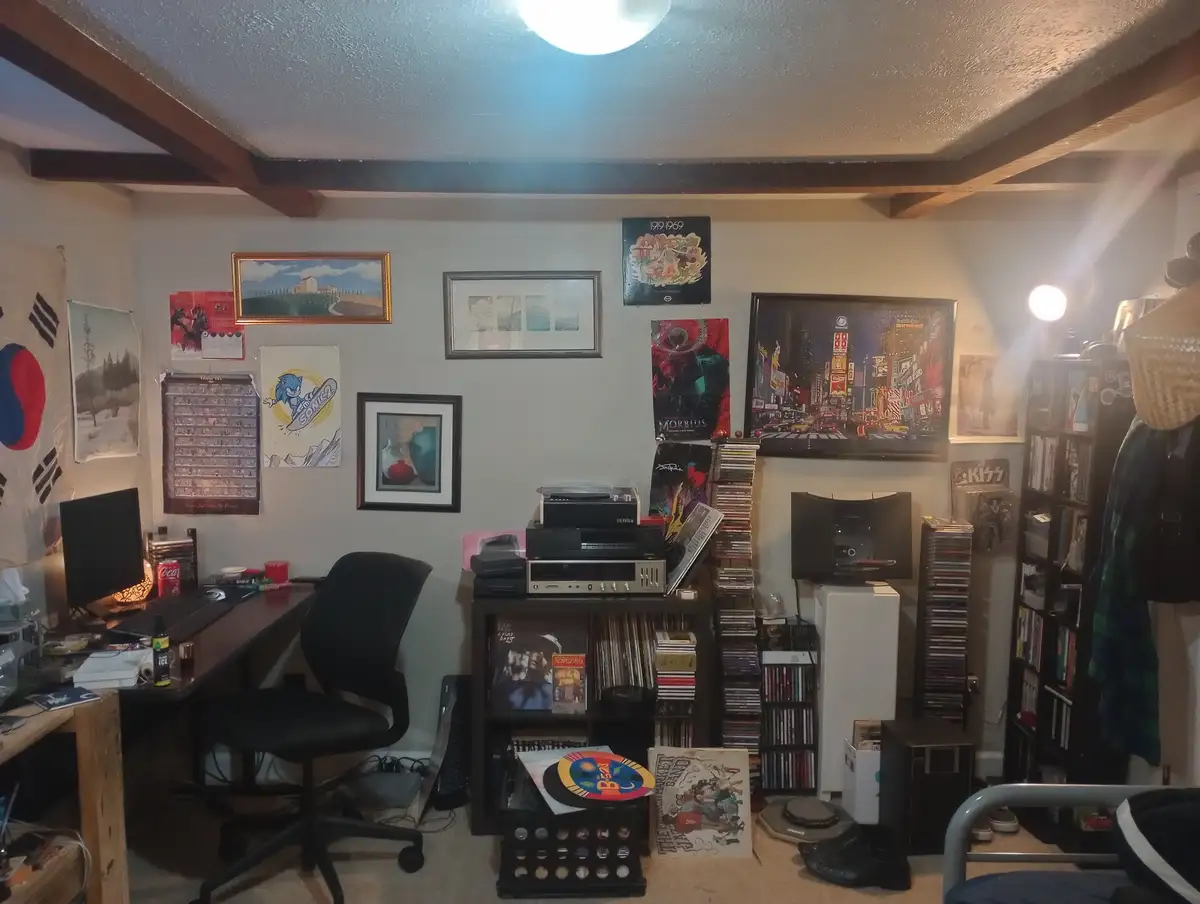 A cluttered room filled with posters, framed art, a desk with a computer, stacks of CDs and vinyl records, and various…