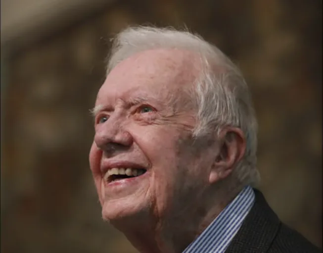 a close-up photo of an elderly white man with white hair, wearing a suit and striped shirt, smiling broadly while looking…