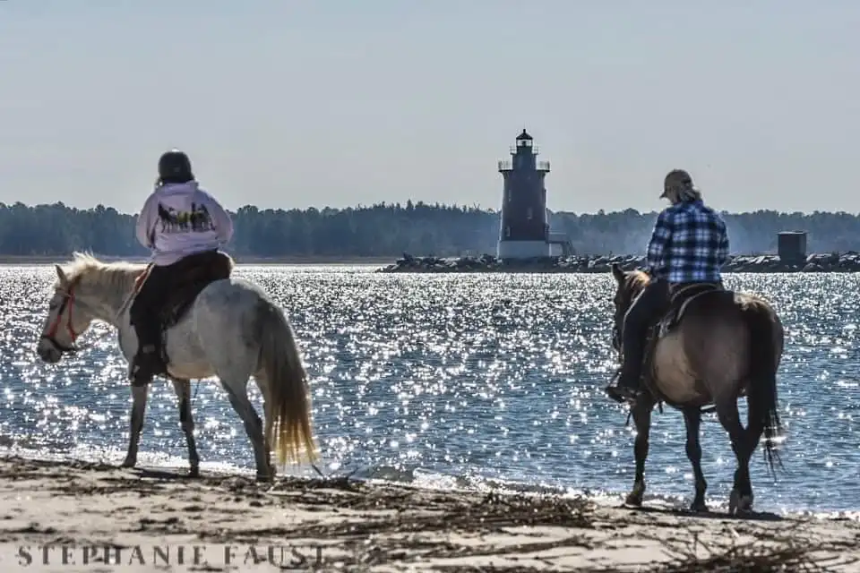 Two people ride horses along a sunlit beach, facing away from the camera toward a lighthouse in the distance. The person on…
