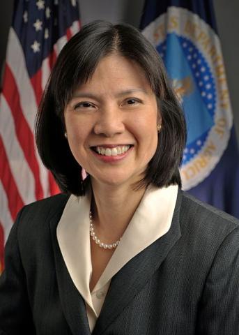 A formal portrait of a smiling woman with dark hair, wearing a dark suit and pearl necklace, standing in front of an…