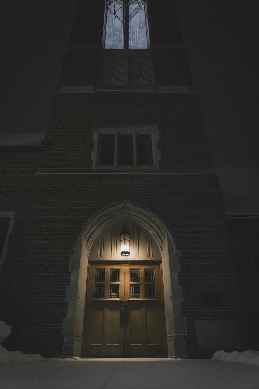 A dark, atmospheric photo of a church entrance at night, featuring a wooden double door under a stone archway, illuminated…