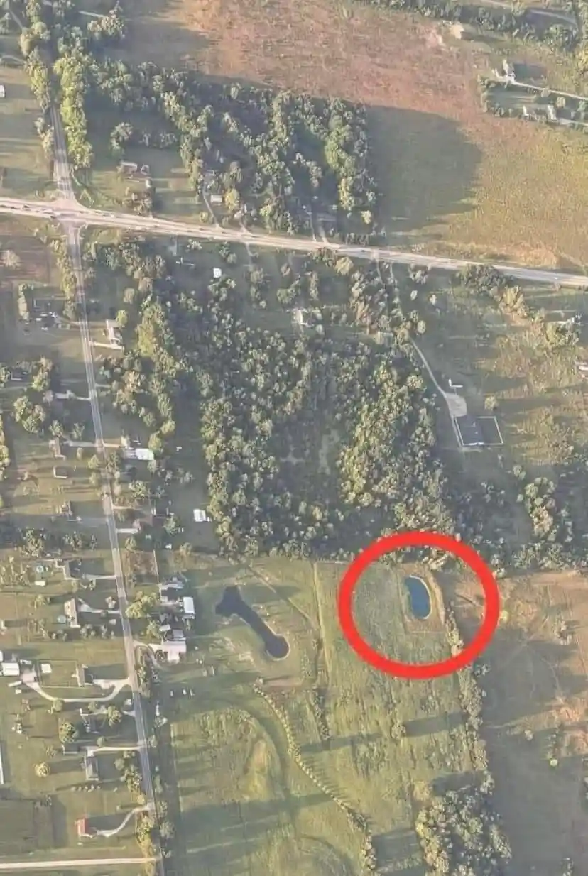 An aerial photograph shows a rural landscape with fields, trees, and roads. A red circle highlights a small, irregularly…