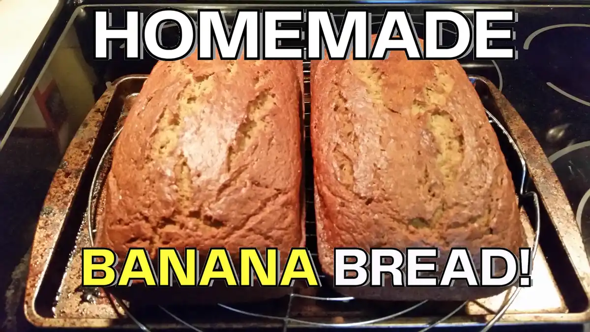 Two loaves of banana bread sit side-by-side on a cooling rack inside a baking pan, resting on a stovetop. Large white text…
