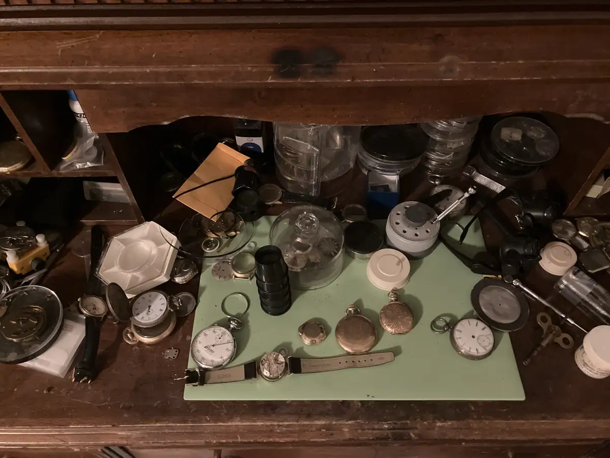 An overhead shot of a cluttered wooden workbench filled with watch parts, tools, and various disassembled timepieces…