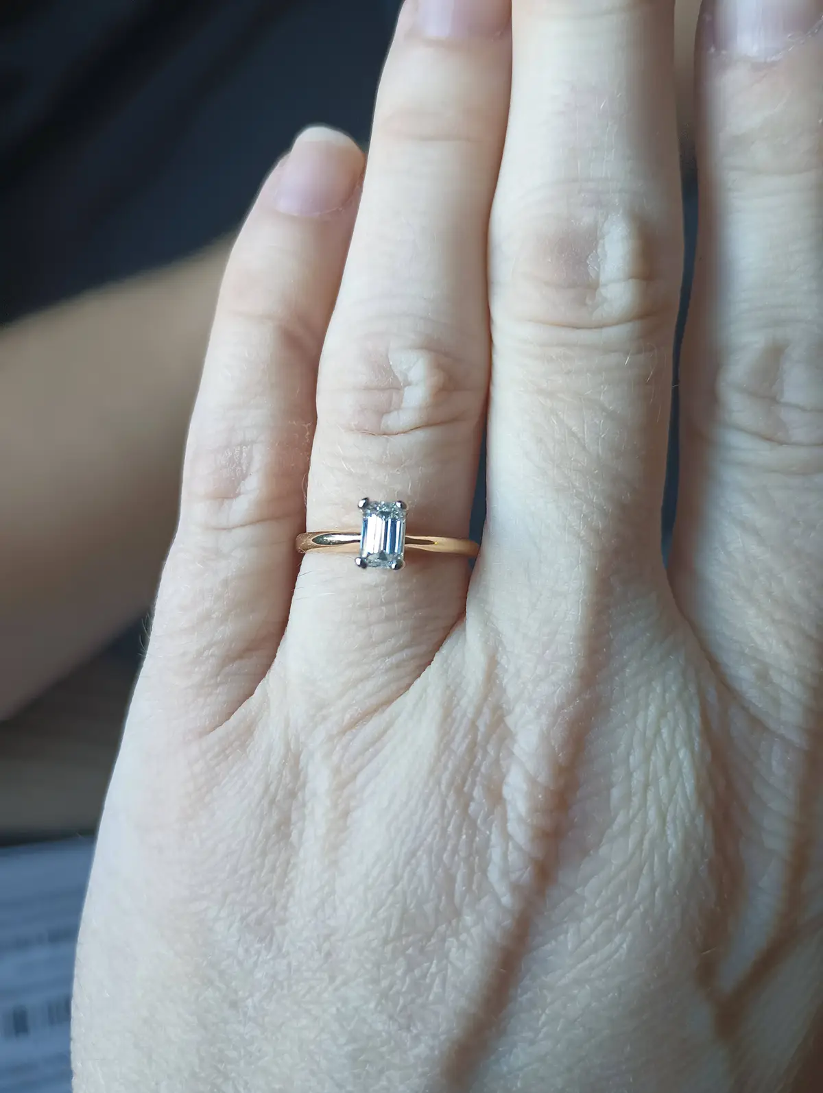 A close-up photo shows a person's hand with a rose gold engagement ring featuring a rectangular emerald-cut diamond on the…