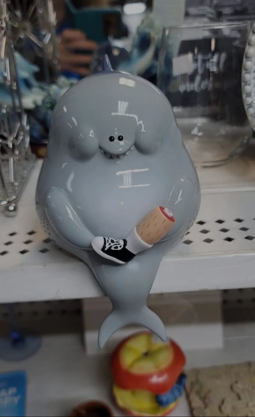 A gray ceramic narwhal figurine sits on a store shelf, drawn with a white chalk-like outline of a top hat, mustache, and a…