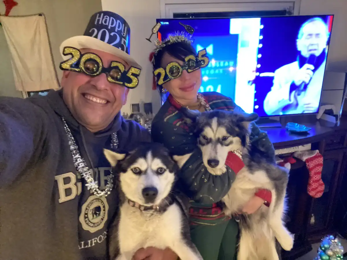 A man and woman in a living room, both wearing glittery '2025' novelty glasses and New Year's hats, pose with two husky dogs…