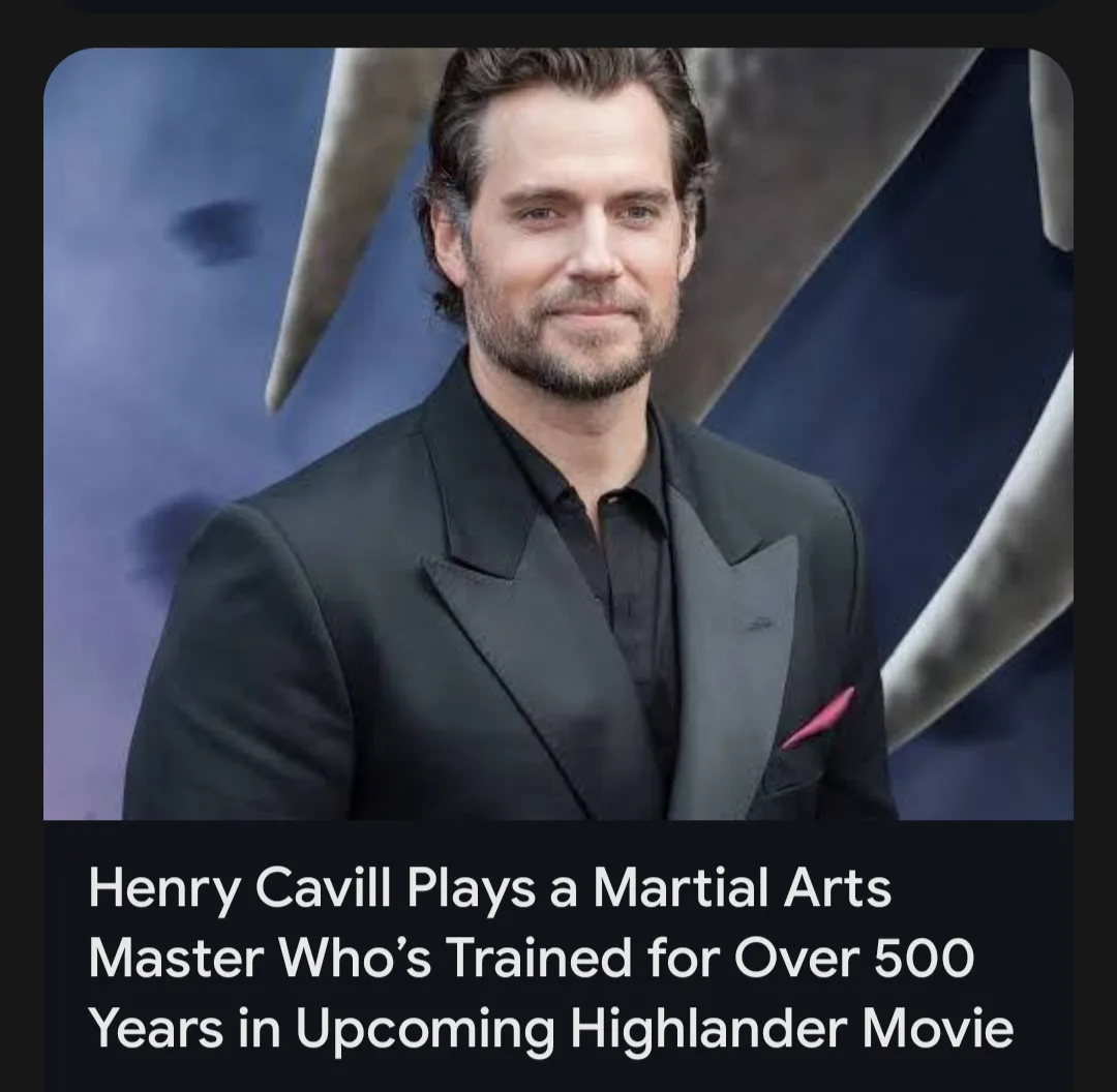 A photo of actor Henry Cavill in a black suit with a pink pocket square, standing in front of a dark blue backdrop with…