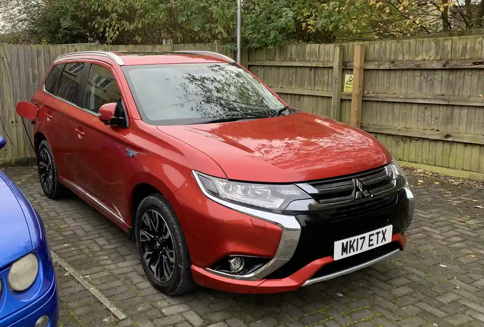 A red Mitsubishi Outlander PHEV is parked on a brick driveway next to a wooden fence, with a blue car partially visible to…