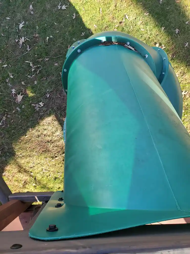 A close-up, top-down view of the top opening of a green plastic playground slide, showing bolts and some dirt or leaves at…