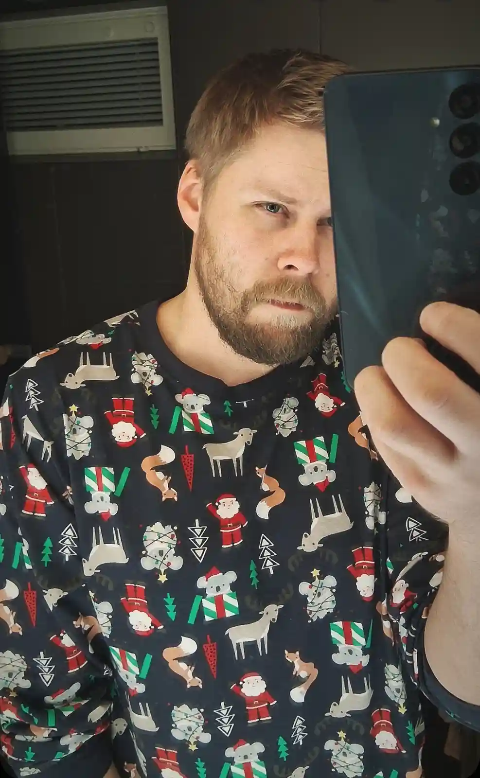 A man with a beard takes a mirror selfie holding a smartphone, wearing a dark shirt covered in repeating cartoon…