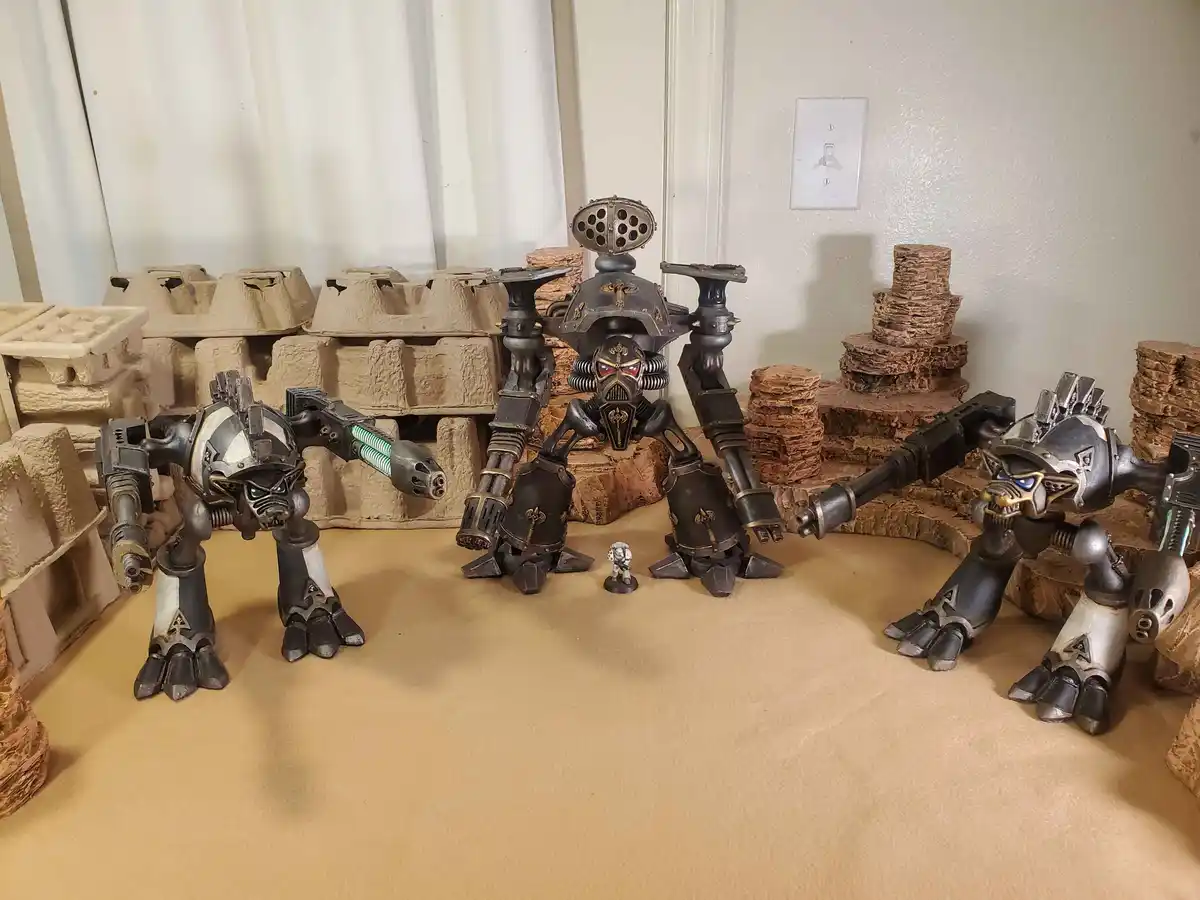 Three large, heavily armed, custom-painted sci-fi mechs stand in a defensive formation around a tiny miniature soldier, with…