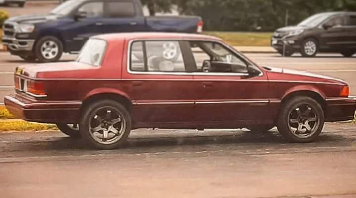 A maroon 1990s sedan with aftermarket black wheels is parked on a street, with other cars visible in the background. The…