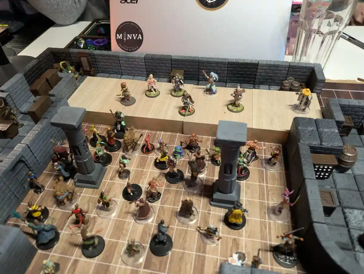 An overhead view of a tabletop roleplaying game setup featuring numerous fantasy miniatures on a wooden grid battlefield…