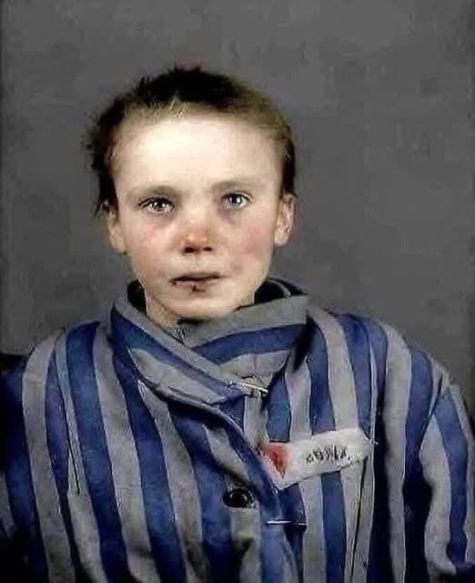 A young girl with pale skin and dark circles under her eyes, wearing a blue and gray striped prisoner uniform with a name…