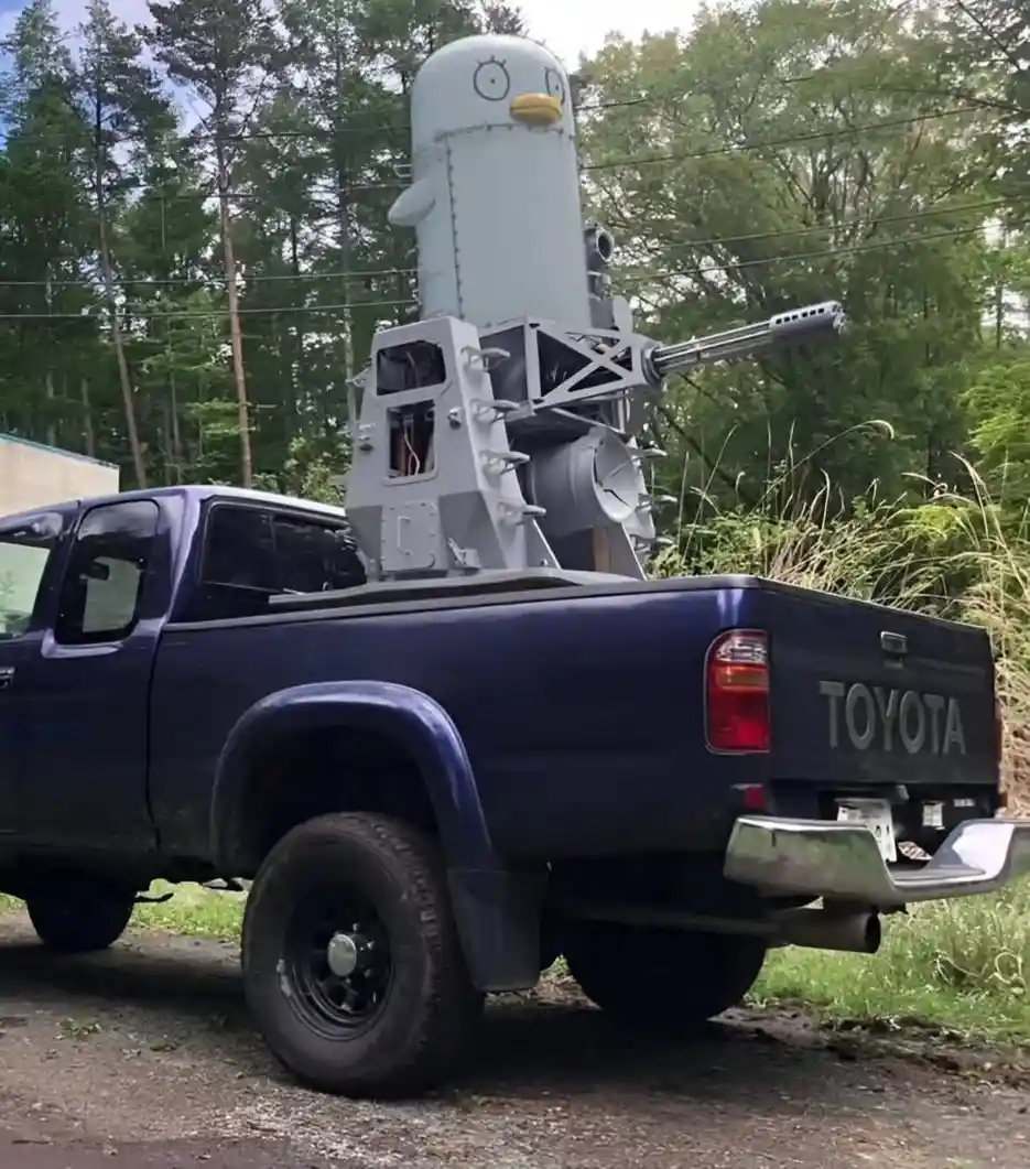 A dark blue Toyota pickup truck has a large, gray, cartoonish penguin-shaped turret mounted in its bed, featuring a yellow…