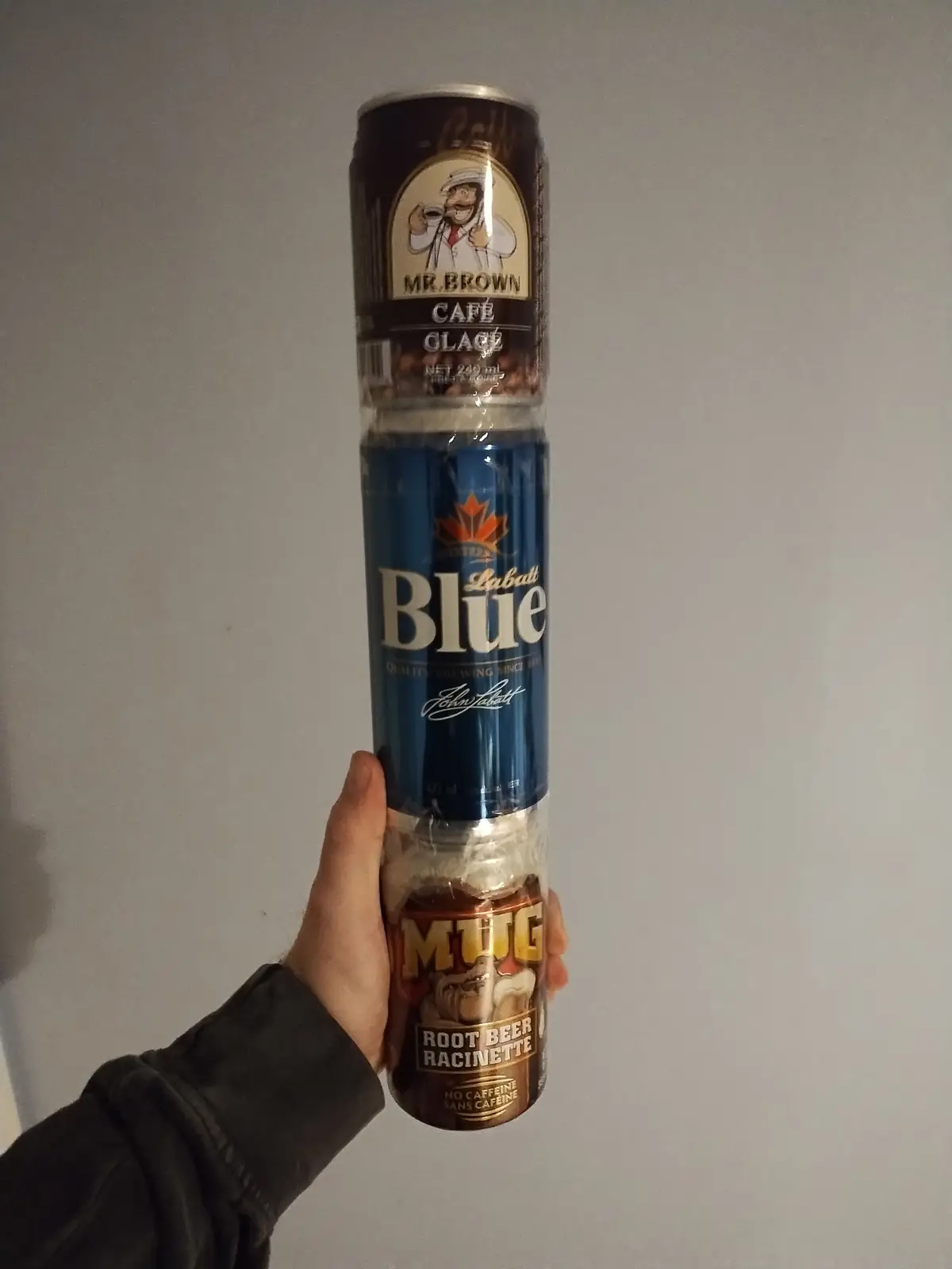 A hand holds three beverage cans stacked vertically in clear plastic wrap: a Mr. Brown Café Glace coffee can on top, a…