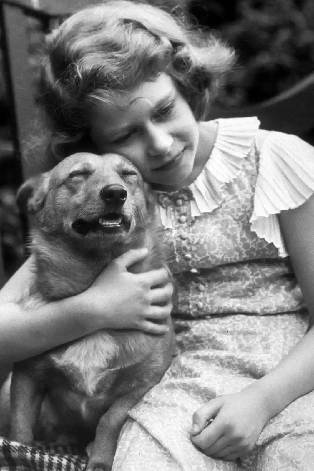 A vintage black-and-white photo shows a young girl in a patterned dress tenderly hugging a dog. The dog has its eyes closed…