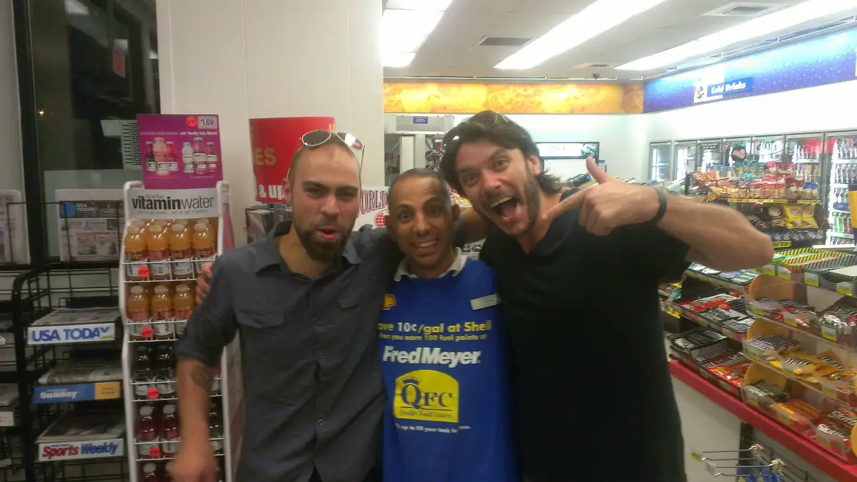 Three men pose together inside a convenience store; the man in the center wears a blue QFC employee shirt, while the man on…
