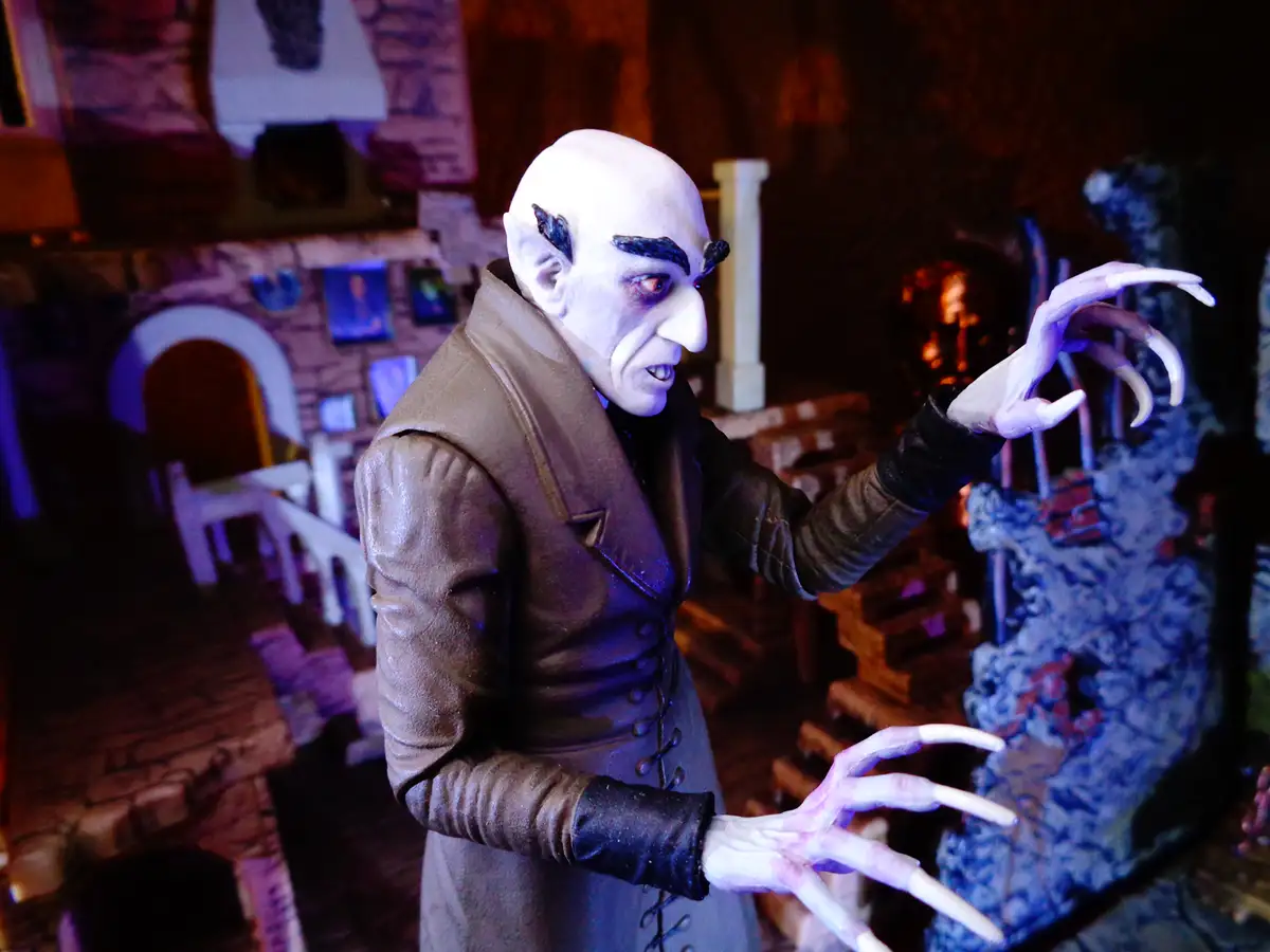 A figurine of a bald, pale vampire with pointed ears and long fingernails, wearing a brown coat and black gloves, stands in…