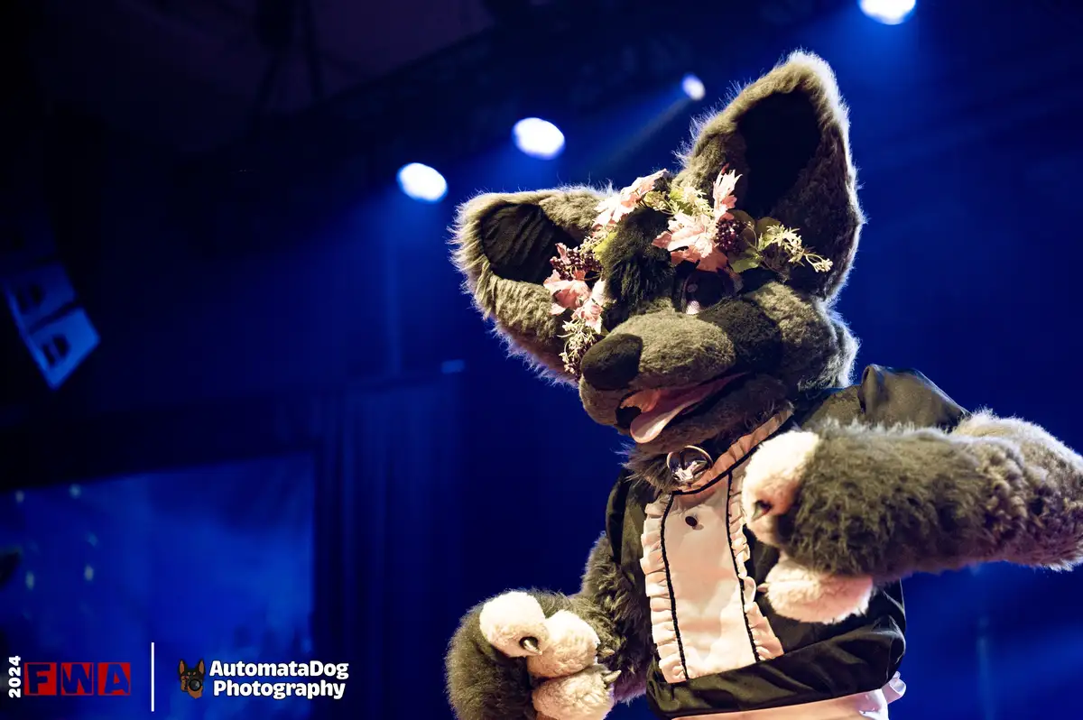 A person in a detailed grey wolf fursuit with large ears, a floral headpiece, and a black and white outfit is posing on a…