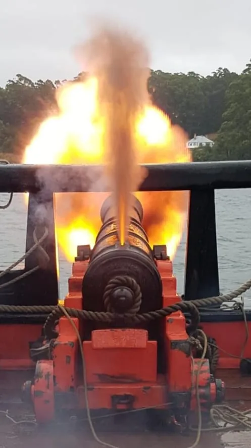 A large, ornate cannon mounted on a red carriage fires a bright, fiery shot, with smoke billowing upward against a backdrop…